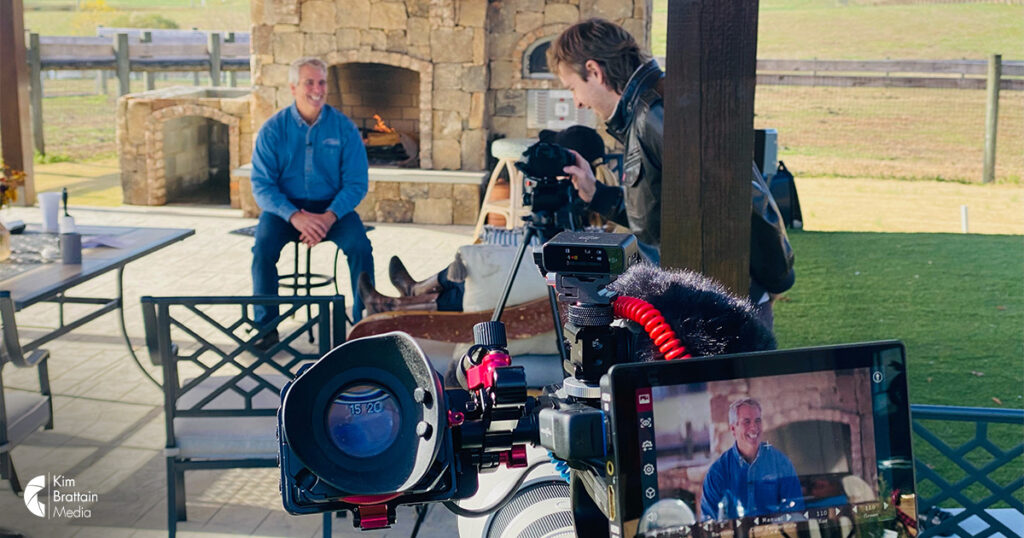 View through a video camera lens capturing a male smiling on a video screen and with Kim Brattain's video production team in Charlotte, NC.